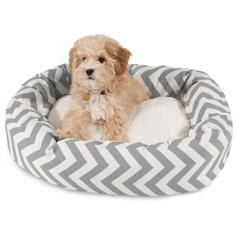 A small, fluffy light brown dog sits in the Majestic Pet Chevron Sherpa Bagel Dog Bed, which features a gray and white chevron pattern and is made of water-resistant outdoor-treated polyester, looking toward the camera.