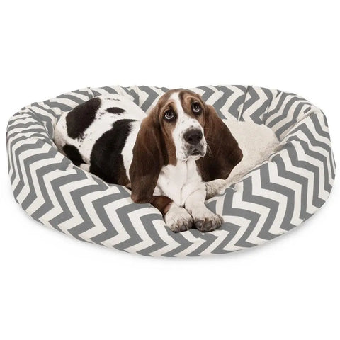 A basset hound with brown, black, and white fur relaxes in the Majestic Pet Chevron Sherpa Bagel Dog Bed, featuring water-resistant outdoor-treated polyester and a gray and white chevron pattern.