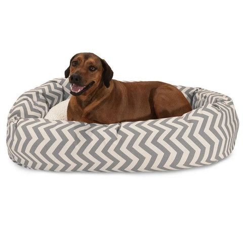 A brown dog relaxes on the Majestic Pet Chevron Sherpa Bagel Dog Bed, featuring a gray and white chevron pattern and water-resistant fabric, gazing at the camera with its mouth slightly open.