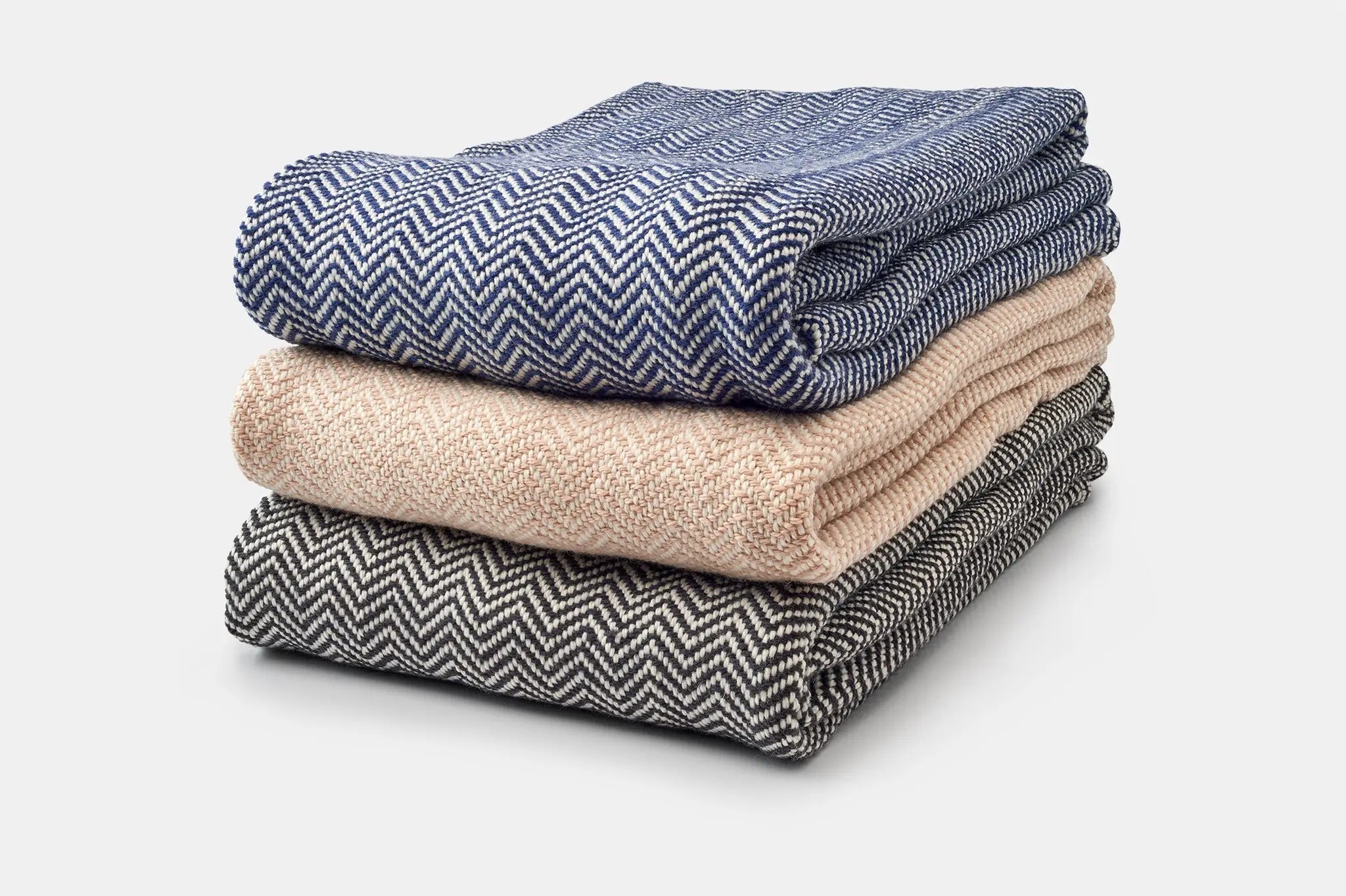 Three folded Chevron Pattern Soft Wool Throw Size Blankets by American Blossom Linens, in blue, beige, and black Climate Beneficial wool, are stacked atop each other against a plain white background.
