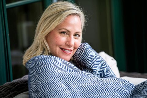 A blonde woman smiles warmly while wrapped in the Chevron Pattern Soft Wool Throw Size Blanket by American Blossom Linens, relaxing indoors near a window.