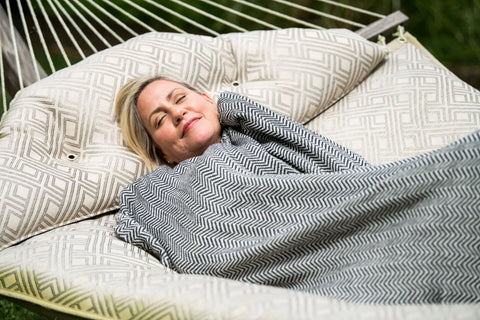 A blonde woman relaxes outdoors on a cushioned hammock, eyes closed in peace, wrapped in the Chevron Pattern Soft Wool Throw Size Blanket by American Blossom Linens.