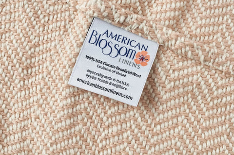 A close-up of the Chevron Pattern Soft Wool Throw Size Blanket by American Blossom Linens shows a label reading: “American Blossom Linens, 100% USA Climate Beneficial Wool, Impeccably made in the USA, americanblossomlinens.com.”.