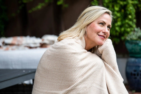 A smiling woman with blonde hair sits outdoors, wrapped in the American Blossom Linens Chevron Pattern Soft Wool Throw Size Blanket. She looks relaxed and content, with greenery and a blurred outdoor bed behind her.