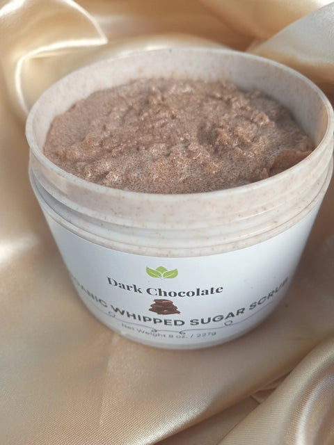 An open jar of Forev'Herbs Chocolate Delight Organic Whipped Sugar Scrub sits on smooth, light beige satin, revealing its fluffy brown texture in a white container with a partially visible label.