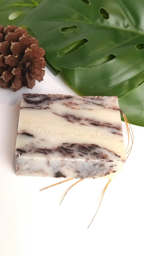 The Forev'Herbs Chocolate Marble Handmade Soap features elegant white and brown swirls and is accented with raffia strands, highlighting its organic, natural appeal. It’s beautifully displayed near a pine cone and a large green leaf.