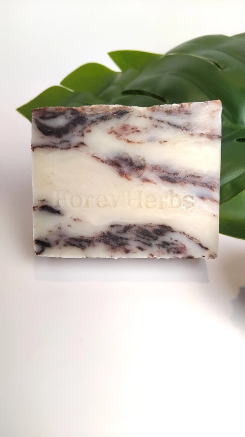 A rectangular Forev'Herbs Chocolate Marble Handmade Soap bar, embossed with the brand name, is placed before green leaves on a white background.