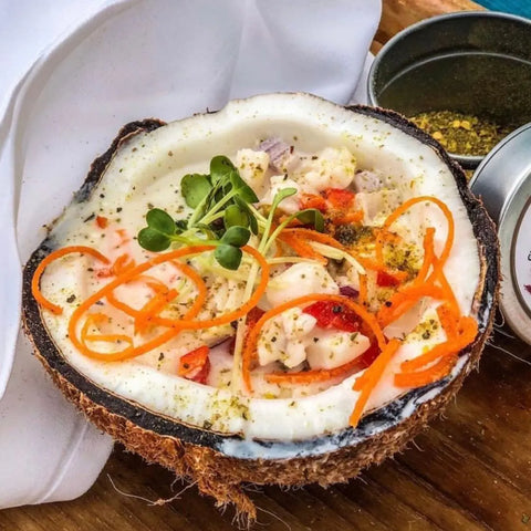 A coconut half with vibrant ceviche—fish, peppers, onions, microgreens, and carrots—is topped with Gustus Vitaes Chucks Picks 10 Pack Collection authentic gourmet seasonings and spice blends, resting beside the tin on a wooden surface.