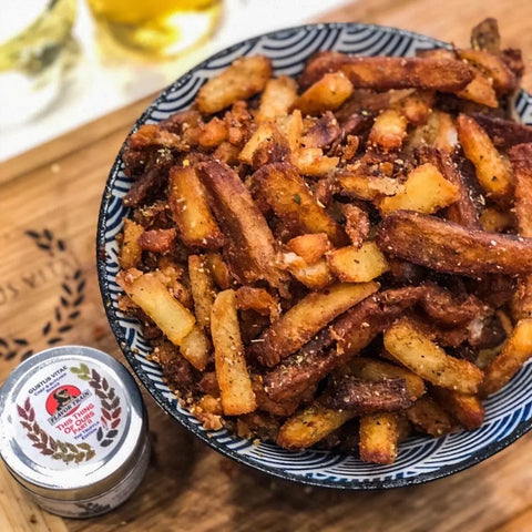 A bowl of crispy French fries topped with Gustus Vitae Chucks Picks Authentic Gourmet Seasonings sits on a board beside a tin of truffle salt and a blurred dish in the background.