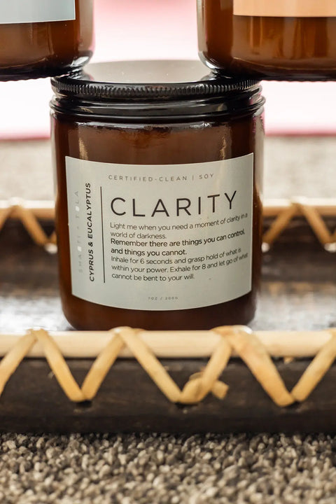 A brown glass Clarity – Cypress & Eucalyptus Soy Candle by Shanti + Tula rests on a wooden tray, its label featuring a relaxation ritual and guidance for finding clarity and letting go. Clean-burning, USA-made wellness candle.