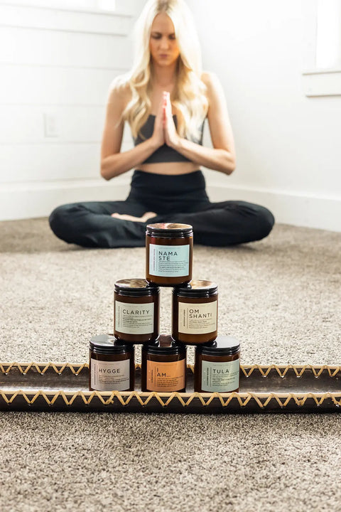 A woman meditates cross-legged on a carpet, while six Shanti + Tula Clarity – Cypress & Eucalyptus Soy Candles are stacked in a pyramid, inviting relaxation with their clean-burning, USA-made wellness ritual fragrance.