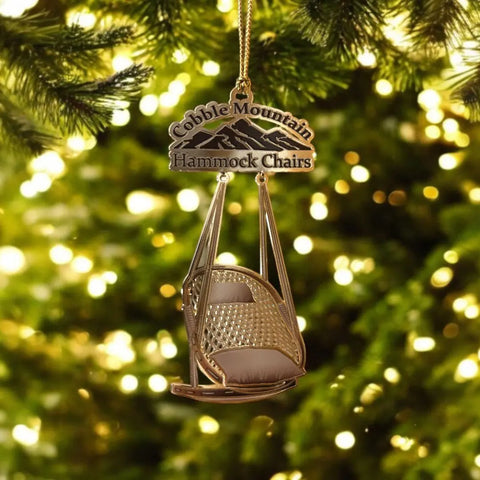 The Cobble Mountain Keepsake Ornament - 1st Edition from Cobble Mountain is a miniature hammock chair and sign, hanging from a branch with blurred golden holiday lights—ideal for adding unique charm to your holiday décor.