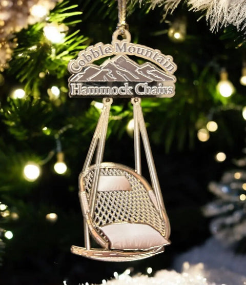 The Cobble Mountain Keepsake Ornament - 1st Edition by Cobble Mountain is a handcrafted American ornament shaped like a hanging chair, featuring Cobble Mountain Hammock Chairs with green pine branches and glowing white holiday accents.