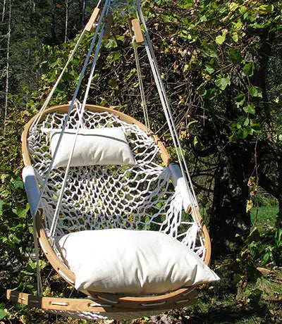 The Cobble Mountain Original Hammock Chair features a wooden frame, white netting, and two white cushions, hanging outdoors among greenery—ideal for relaxed outdoor living.