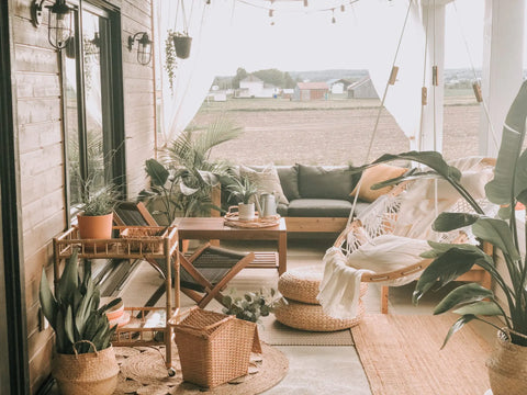 A cozy patio with a sofa, Cobble Mountains Original Hammock Chair, and wooden furniture, decorated with potted plants, cushions, woven baskets, and string lights—perfect for outdoor living overlooking fields and distant houses.