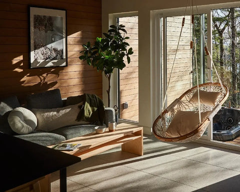 A cozy living room with wood-paneled walls, a gray sofa, wooden coffee table, potted plant, and a Cobble Mountain Original Hammock Chair by large windows that overlook trees and an outdoor hot tub as sunlight streams in.