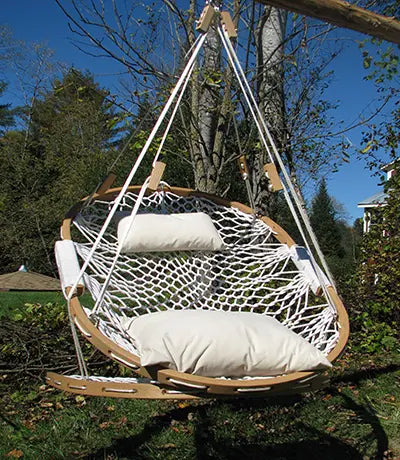 The Summit Hammock Chair by Cobble Mountain features a round wooden frame, white netting, and two white cushions. It hangs from a wooden support, surrounded by grass, trees, and a blue sky.