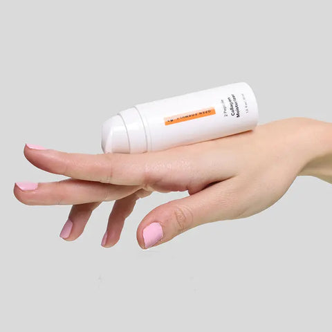 A hand with light pink nail polish holds a white Open Formula Collagen Peptide Moisturizer bottle featuring an orange label, set against a plain gray background.