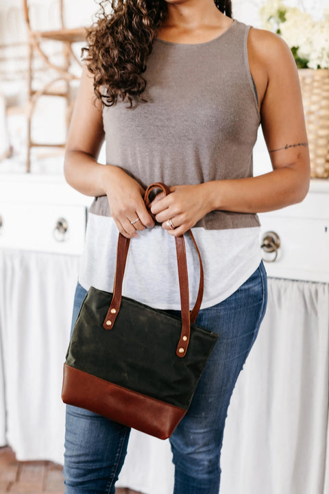 A woman in a sleeveless top and jeans stands indoors, holding the Urban Southern Mini Canvas Tote with both hands in front of her. Her face is not shown.