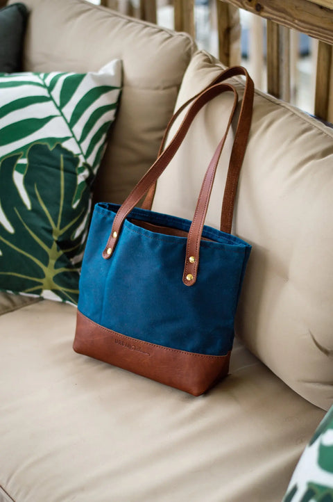 The Urban Southern Mini Canvas Tote, featuring blue and brown colors with leather straps, is displayed on a beige couch next to green and white leaf-patterned pillows.