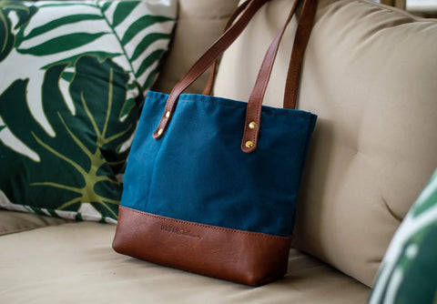 A blue and brown Urban Southern Mini Canvas Tote with two brown handles rests on a beige sofa beside green and white leaf-patterned cushions.