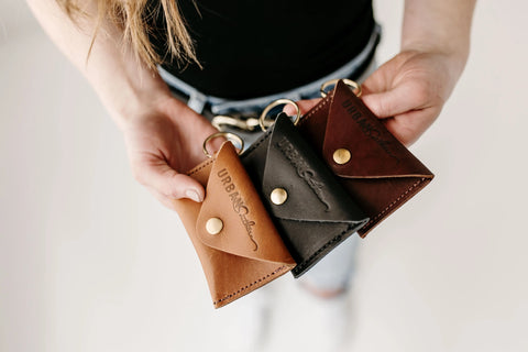 A hand holds three Urban Southern Keychain Coin Purses made of full-grain leather in brown, black, and dark brown, each with a brass button and keyring—stylish essentials embossed with the brand logo.