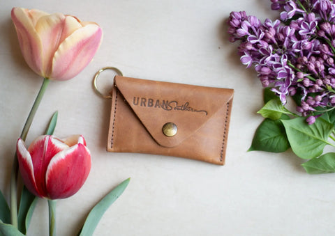 A compact brown Keychain Coin Purse by Urban Southern rests on a light surface, surrounded by pink and red tulips and clusters of purple lilac flowers.