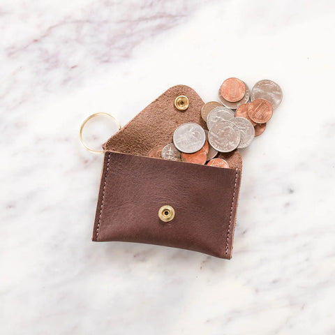 The Urban Southern Keychain Coin Purse, crafted from full-grain leather with a gold snap button, sits partially open as assorted coins spill onto a white marble surface.