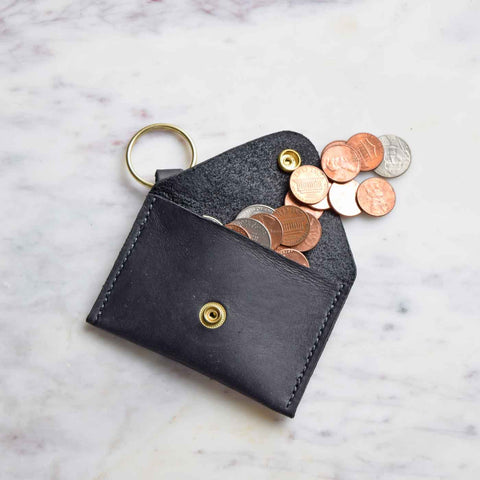 The Urban Southern Keychain Coin Purse, a compact black full-grain leather pouch with a snap button and keyring, lies open on a white marble surface with several U.S. coins spilling out.