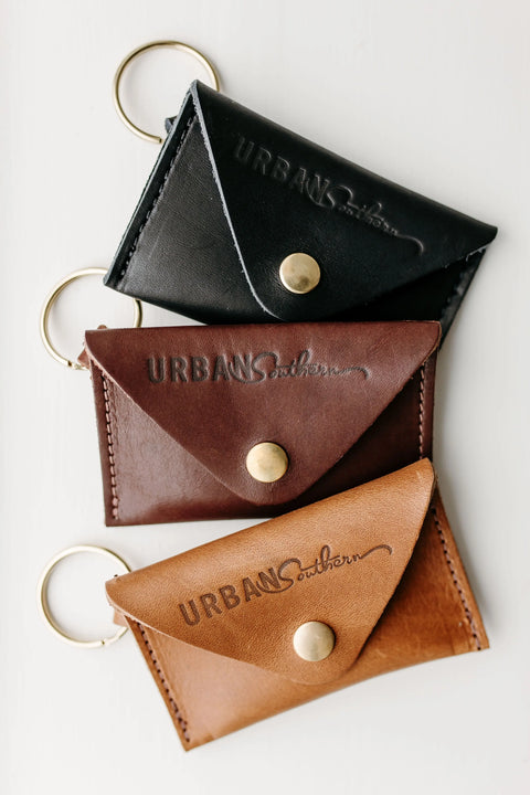Three Urban Southern Keychain Coin Purses in black, dark brown, and tan full-grain leather with brass buttons and keyrings are displayed vertically on a white background. Each purse is embossed with the brand name.