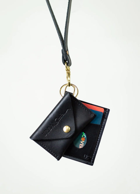 The Urban Southern Keychain Coin Purse, made of black full-grain leather with a snap button, card holder, and gold keyring, hangs from a black lanyard. Two colorful cards are inside, displayed against a plain white background.