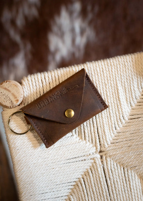 A brown full-grain leather Keychain Coin Purse by Urban Southern, featuring a brass button and keyring, rests on a woven cream textile. A wooden tag is attached to the keychain coin purse.