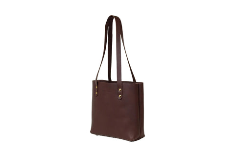 The Urban Southern Mini Urban Tote is a brown luxury leather bag with two long shoulder straps and brass rivets, standing upright against a white background—an American-made handbag crafted for timeless style.