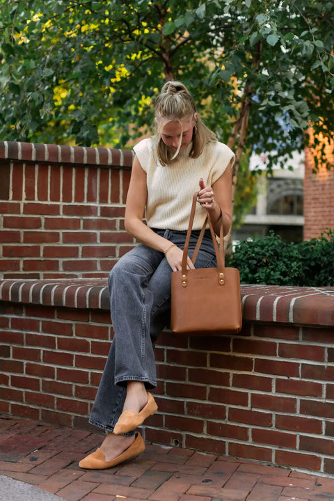 A woman sits on a brick wall outdoors, adjusting her tan shoe. She wears a sleeveless beige sweater, gray jeans, and carries the Urban Southern Mini Urban Tote. Green foliage and additional brick walls are in the background.