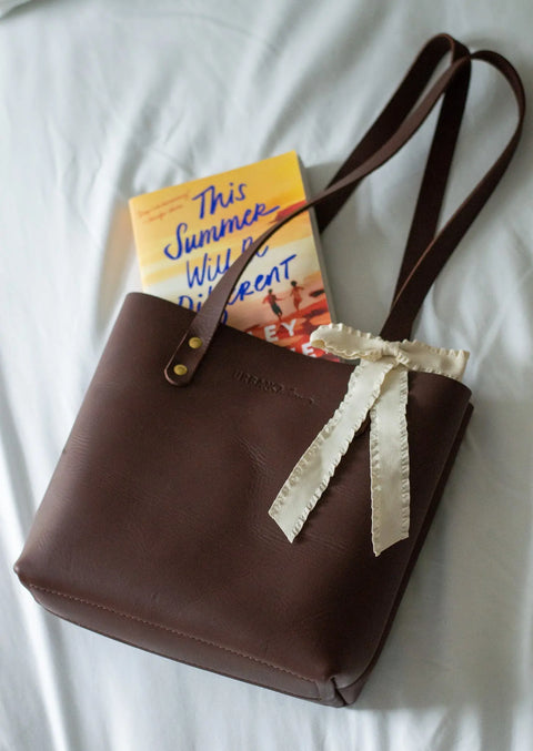 A Mini Urban Tote by Urban Southern, in brown leather with a cream ribbon bow on the handle, sits on a white surface with the book This Summer Will Be Different partially tucked inside.