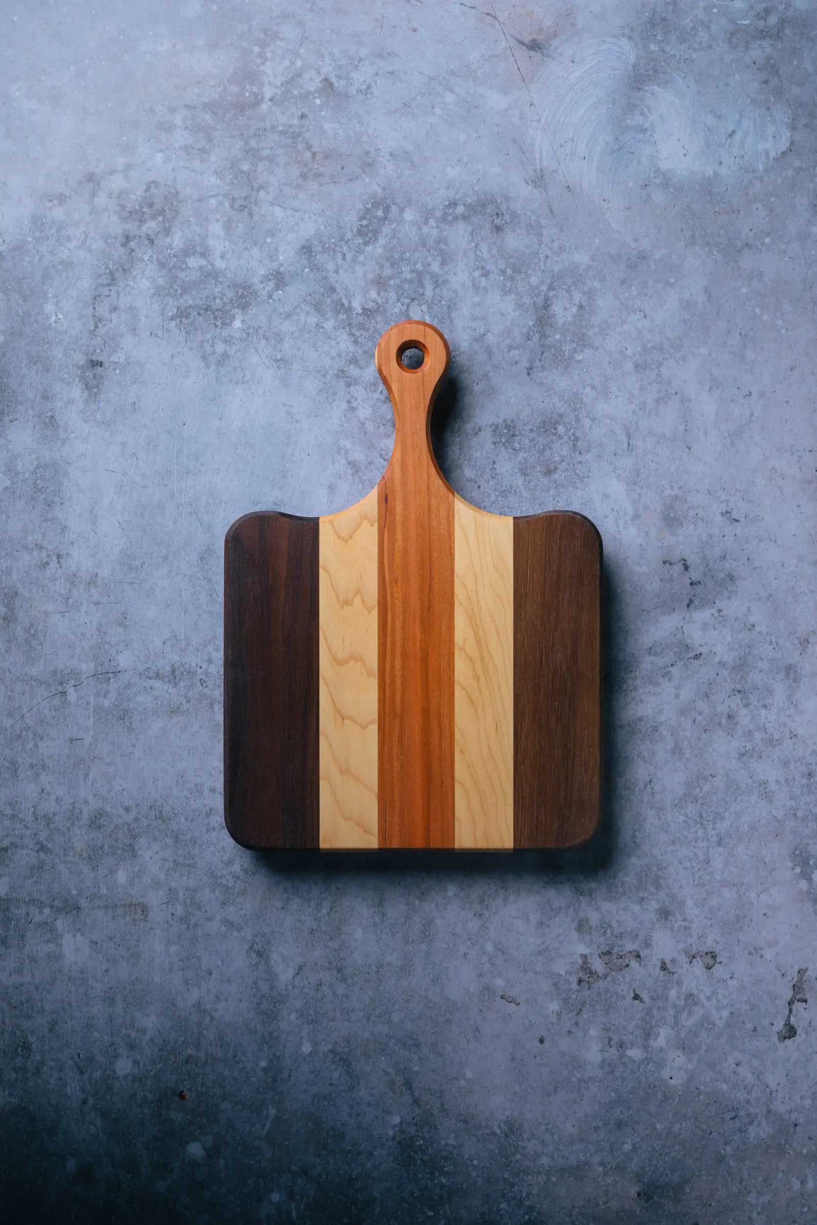 The Bread Board - Compact by Montana Block, an American-made rectangular cutting board with a handle and alternating light and dark wood stripes, rests on a textured gray surface.