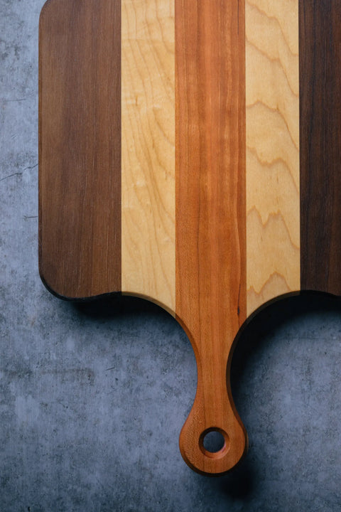 The Bread Board - Compact by Montana Block, made of solid wood with alternating dark and light stripes and a rounded handle with hole, rests on a gray textured surface. Only part of this stylish bread board is visible in the image.