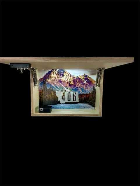 The Regular Size Concealment Cabinet by B. James Design & Woodwork features a wooden shelf, which when flipped reveals a mountain landscape with trees, a lake, and “406” at the center, all on a black background. The interior is fully customizable.