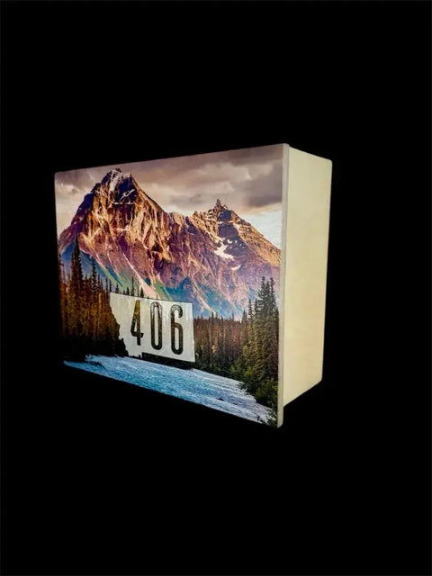 The Regular Size Concealment Cabinet by B. James Design & Woodwork is a wooden block featuring a river, pine trees, and snow-capped mountain photo, with street number 406 on the front against a black background for added security.