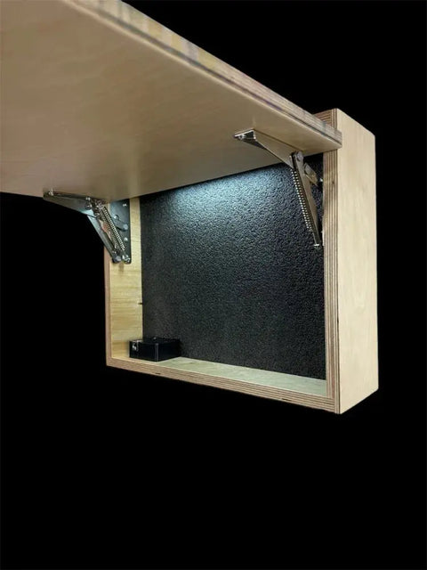 The Regular Size Concealment Cabinet by B. James Design & Woodwork is shown open under a table, featuring a customizable interior with a black textured back, LED lighting, and a small black device, all set against a completely black background.