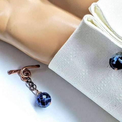 A close-up of a persons arm in a white textured French cuff shirt, fastened with Alexa Martha Designs’ Copper Child Abuse Prevention Awareness Chain Cufflinks featuring handmade copper and dark blue marbled beads.
