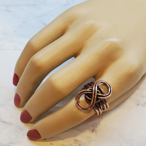 A mannequin hand with red nails showcases the Copper Eternity Ring by Alexa Martha Designs on the ring finger, set against a white marble background.