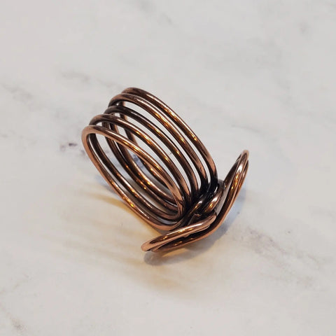 The Copper Eternity Ring by Alexa Martha Designs features handmade overlapping copper coils in a spiral, layered design and is pictured on a white marble surface.