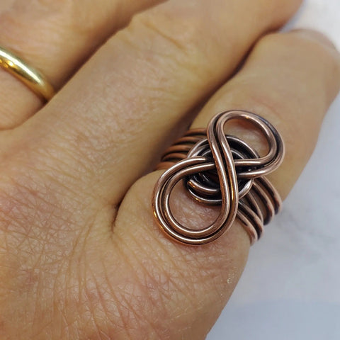 A hand wears the Copper Eternity Ring by Alexa Martha from Alexa Martha Designs, featuring an intricate looping infinity symbol, next to a simple gold band on another finger.