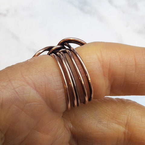 A hand showcases several copper-colored rings, including the Copper Eternity Ring by Alexa Martha from Alexa Martha Designs, stacked on a finger against a softly blurred background.