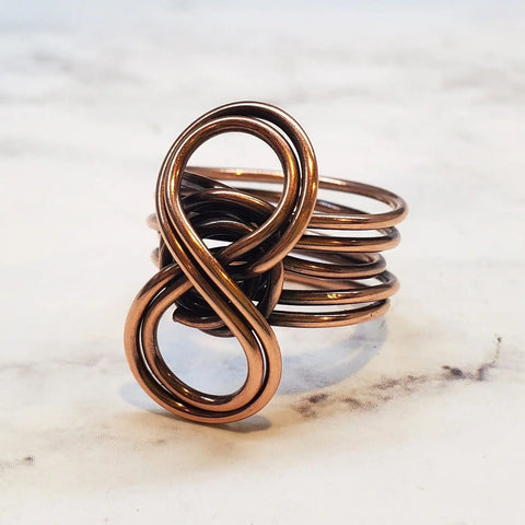 The Copper Eternity Ring by Alexa Martha Designs features an elegant infinity symbol crafted from hand-coiled copper wire, creating multiple bands for a unique, handmade look. The ring is pictured on a white marble surface.