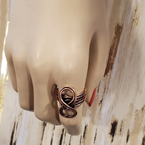 A mannequin hand showcases the Copper Eternity Ring by Alexa Martha from Alexa Martha Designs, featuring a handmade abstract looped design on the middle finger. The background has light tones with brown and gold streaks.