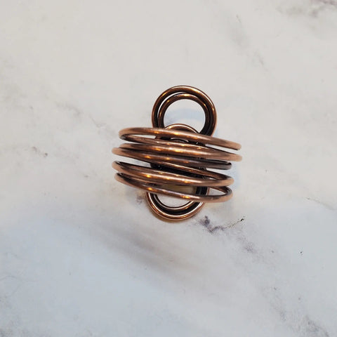 The Copper Eternity Ring by Alexa Martha Designs, featuring multiple interlocking copper loops in an infinity shape, is displayed on a light, marbled surface.