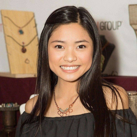 A young woman with long black hair and an off-the-shoulder black top smiles at the camera wearing the Alexa Martha Designs As Seen on Ashley Liao Copper Turquoise Wire Wrapped Necklace, with jewelry displays in the background.