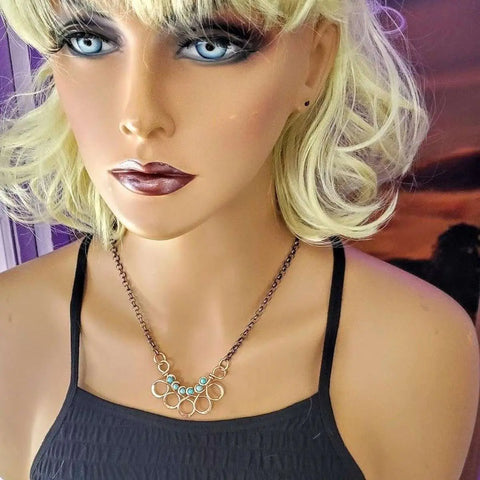 A blonde mannequin with blue eyes and dark makeup wears a black spaghetti strap top and the Alexa Martha Designs As Seen on Ashley Liao Copper Turquoise Wire Wrapped Necklace. The background is purple and orange.